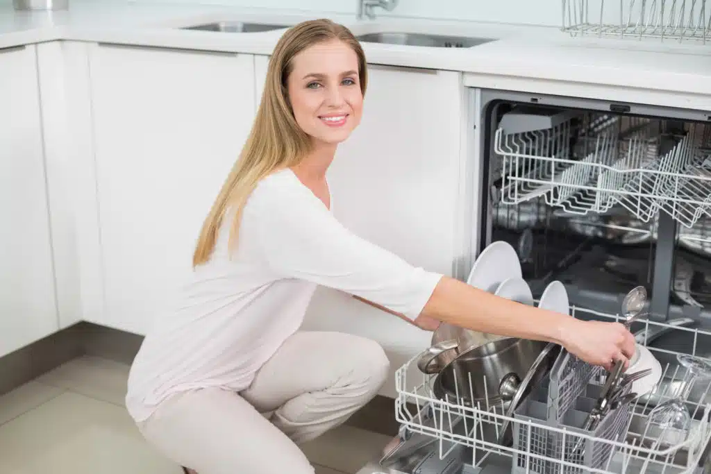 Smiling gorgeous model kneeling next to dish washer in bright kitchen